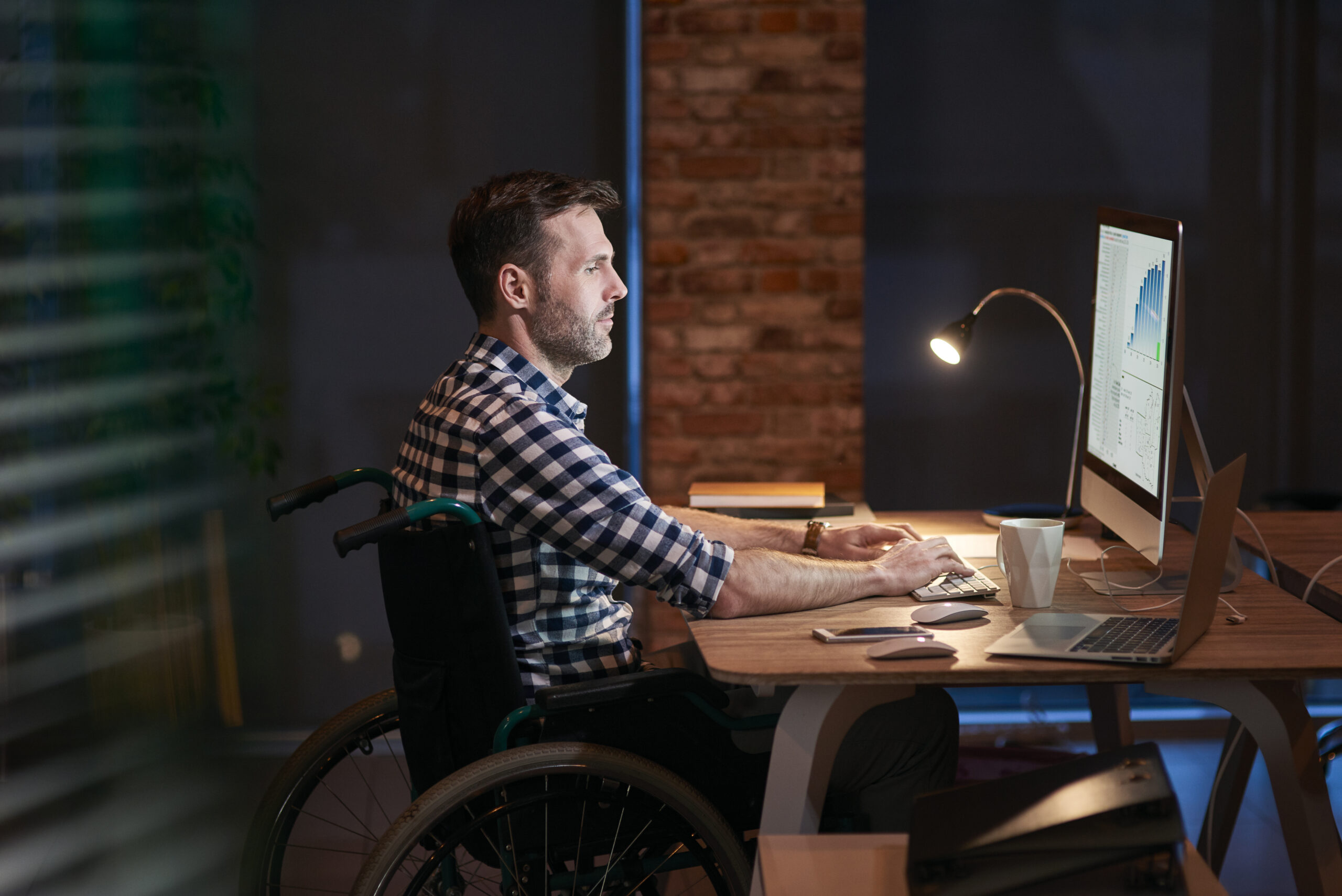 Vue latérale d'un homme handicapé en fauteuil roulant, travaillant à son bureau devant son ordinateur. Il représente les personnes étant affectés par divers handicaps et nécessitant une amélioration de l'accessibilité web, qui est la mission principale d'AccessiWeb.